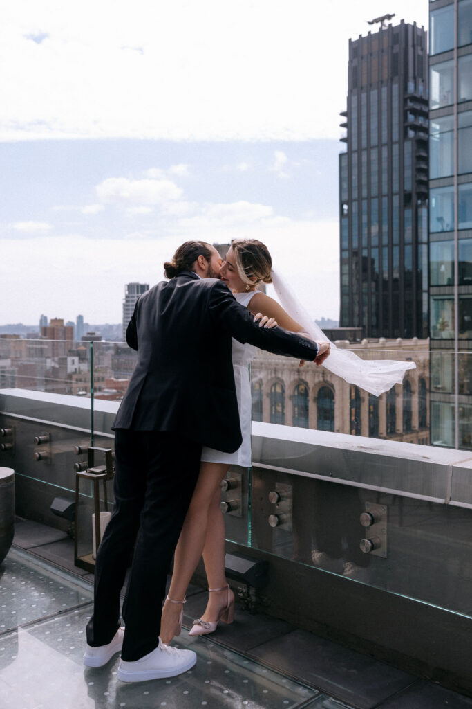 Couple sharing an intimate neck kiss on a rooftop with a city skyline view, capturing a romantic pre-wedding moment