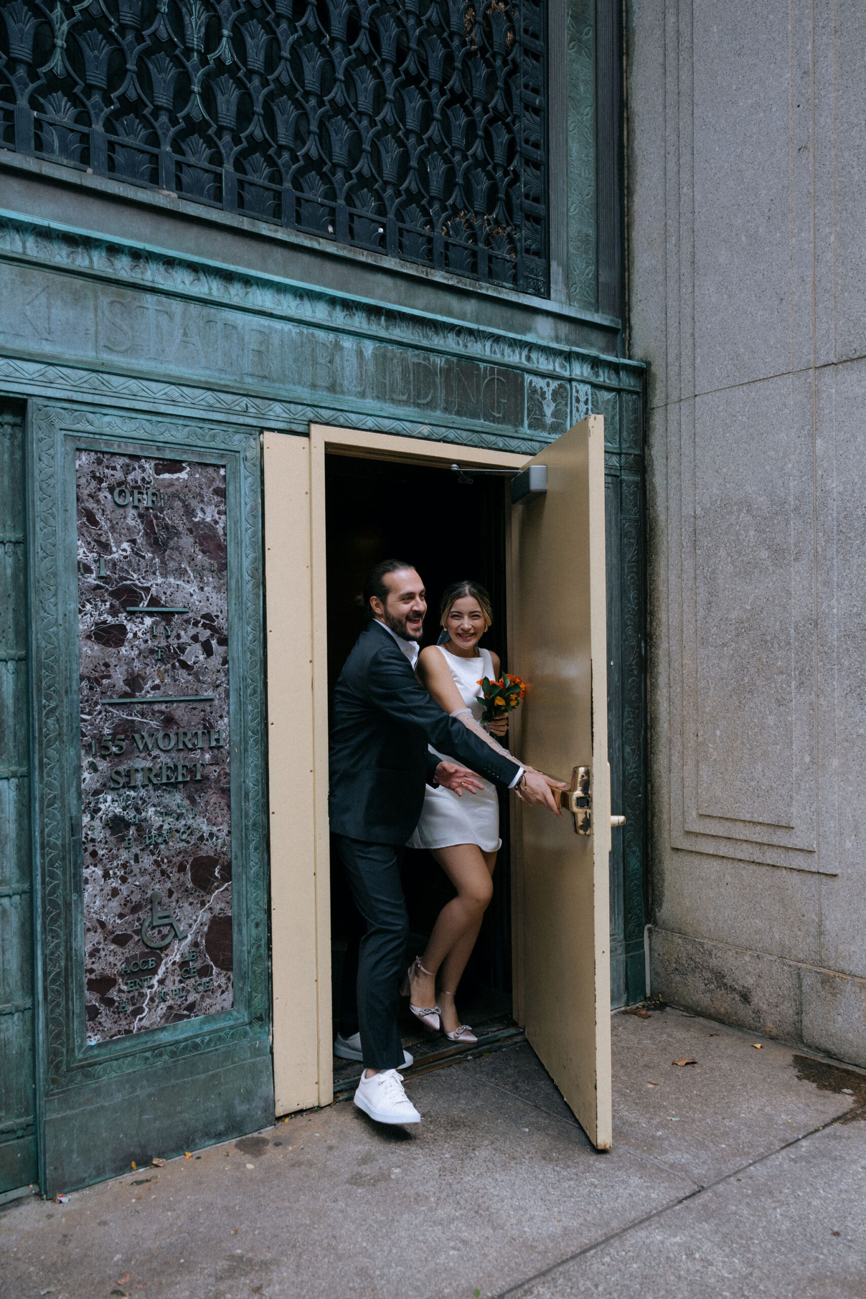 A newlywed couple exiting the City Hall building in NYC. The groom, wearing a black suit and white sneakers, holds the door open with a joyful expression, while the bride, in a short white dress and veil, steps out with a radiant smile, holding a bouquet of orange flowers. The ornate architectural details of the entrance serve as a backdrop to their celebratory moment.