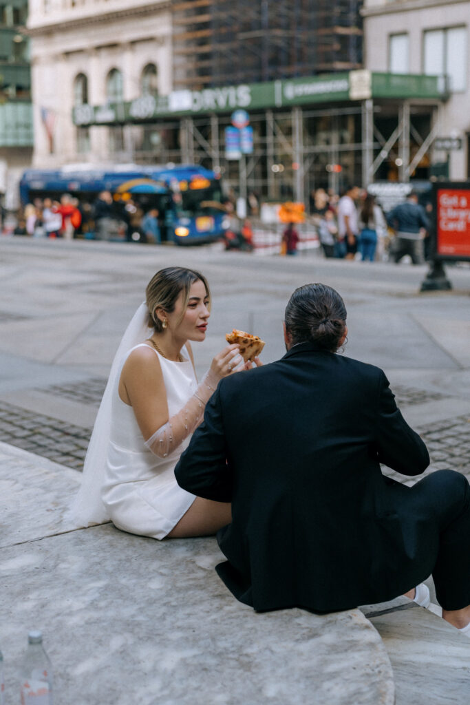 Couple sitting by the New York Public Library, happily sharing pizza as they celebrate their recent wedding in NYC.