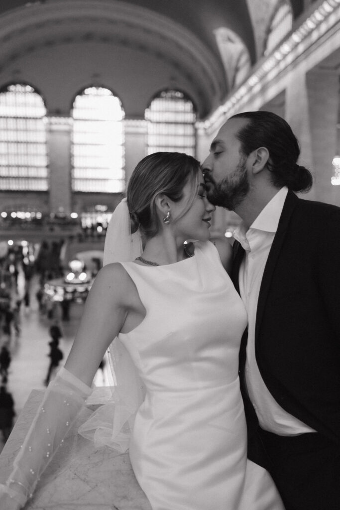 Couple joyfully capturing their elopement at Grand Central Terminal, with the iconic clock and bustling crowd creating a lively NYC backdrop.