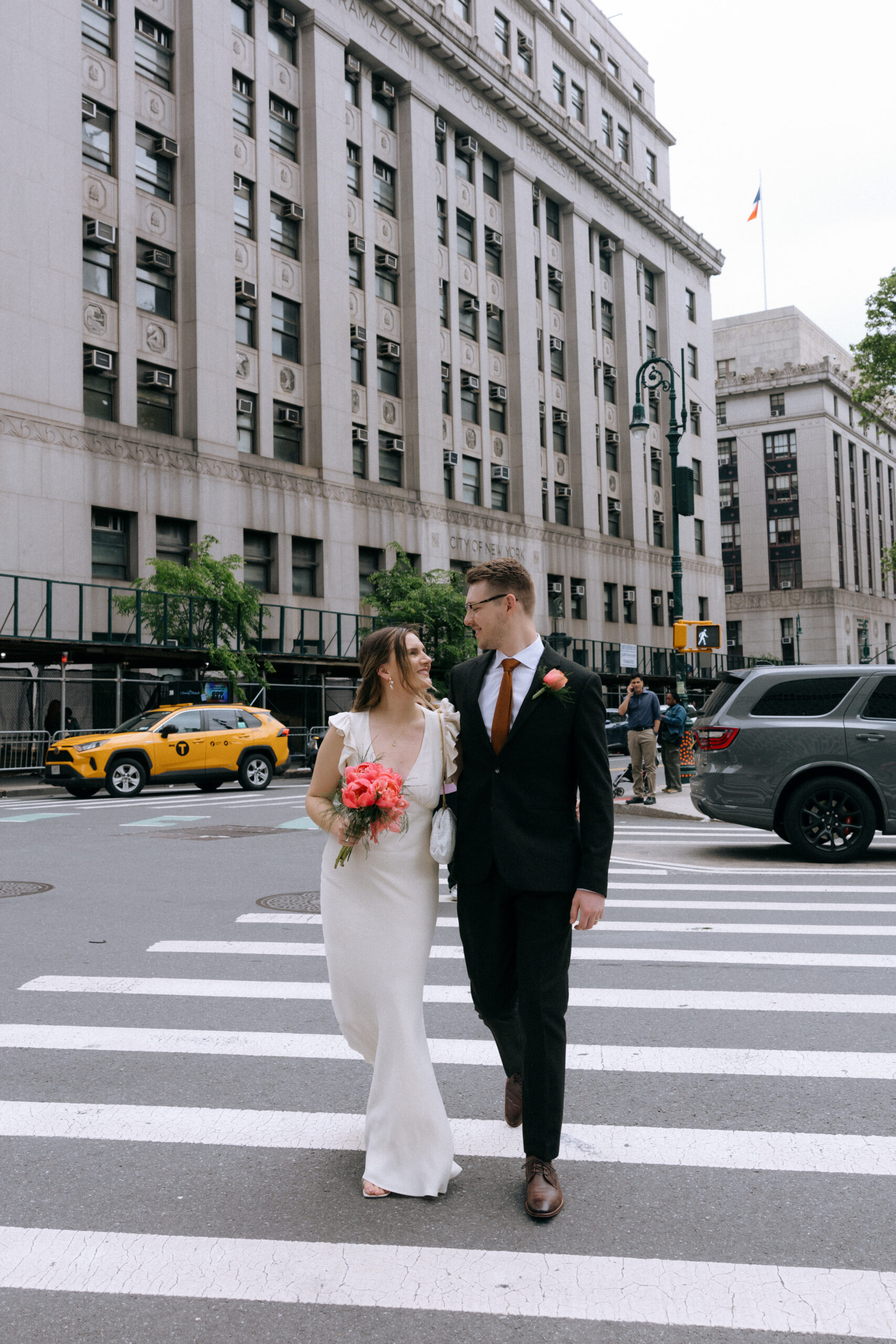 A newlywed couple walking arm-in-arm across a crosswalk in front of a NYC municipal building.