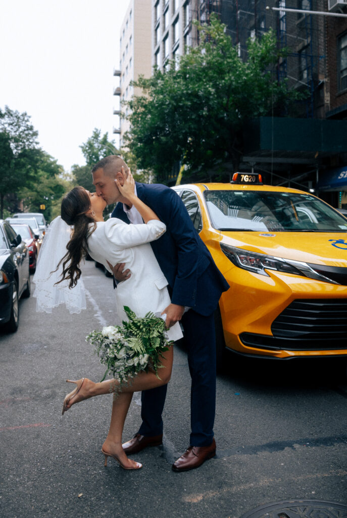 A bride in a white blazer dress and veil kisses her groom, dressed in a blue suit, on a New York City street with a yellow taxi in the background.