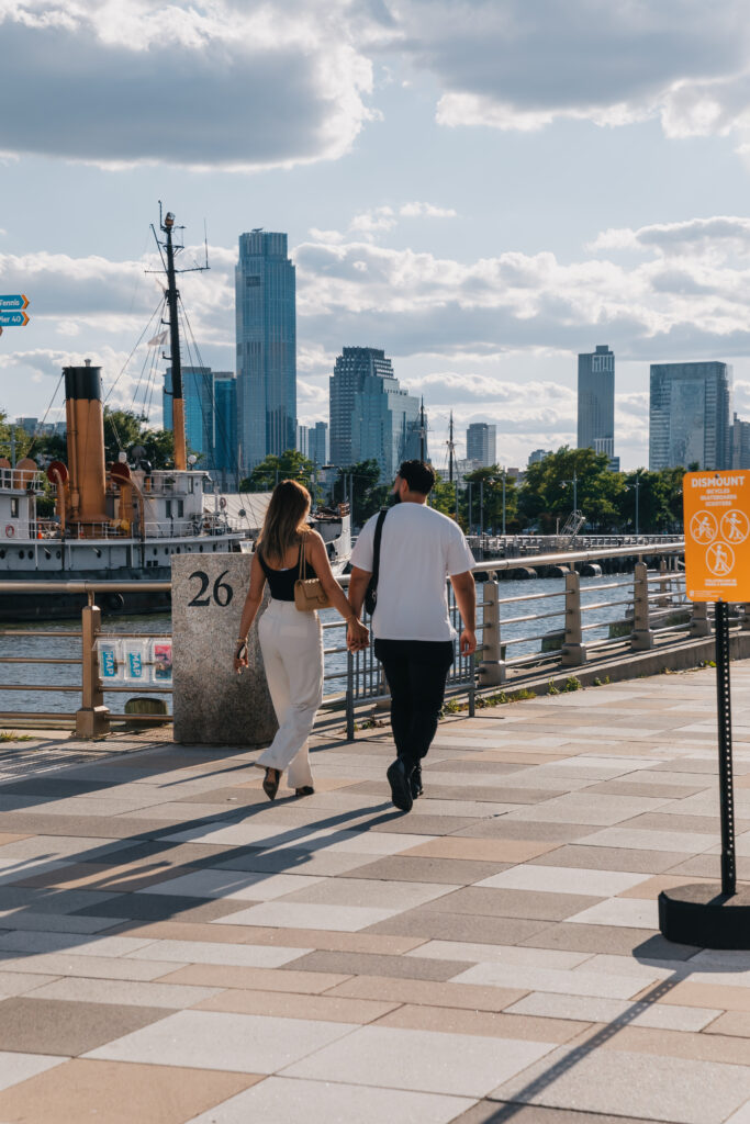 Couple walking hand in hand along the NYC waterfront with the skyline in the background.