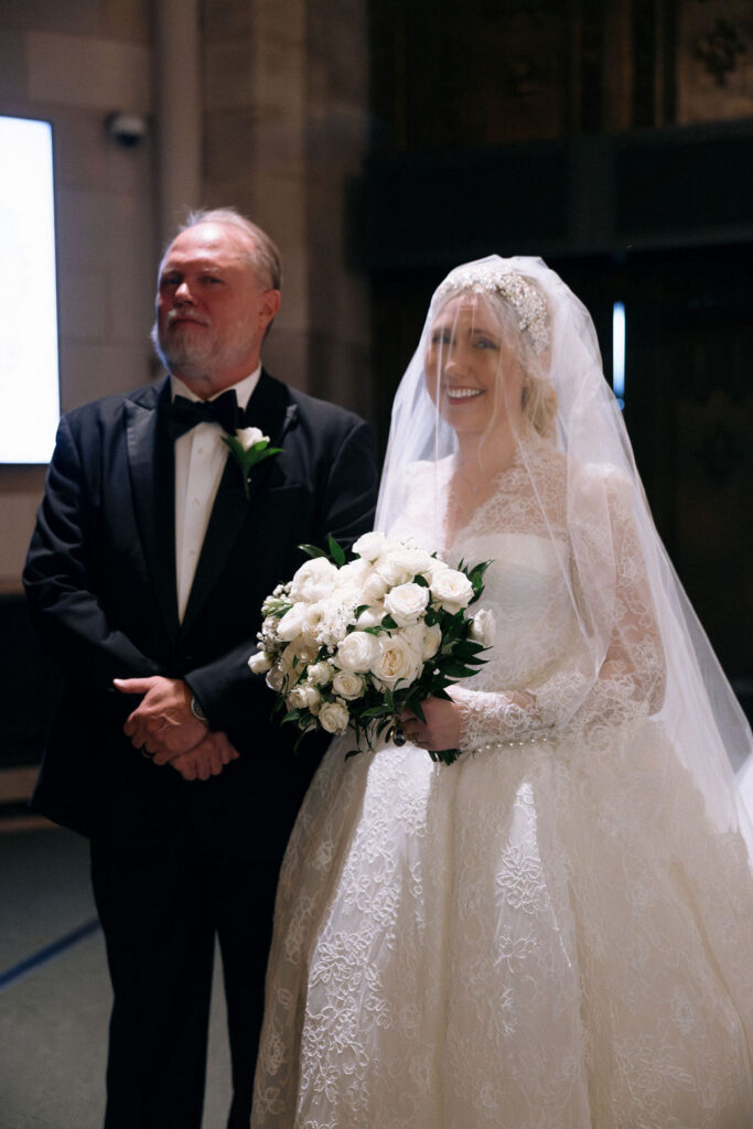 A radiant bride in a luxurious lace gown with a white rose bouquet, standing with her father before walking down the aisle at an elegant NYC wedding.