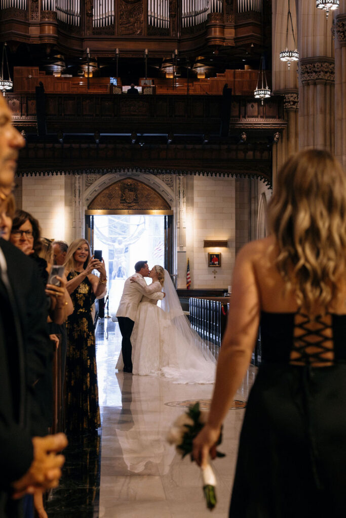 The bride and groom share a celebratory kiss at the end of the aisle, surrounded by joyful guests at their NYC cathedral wedding.