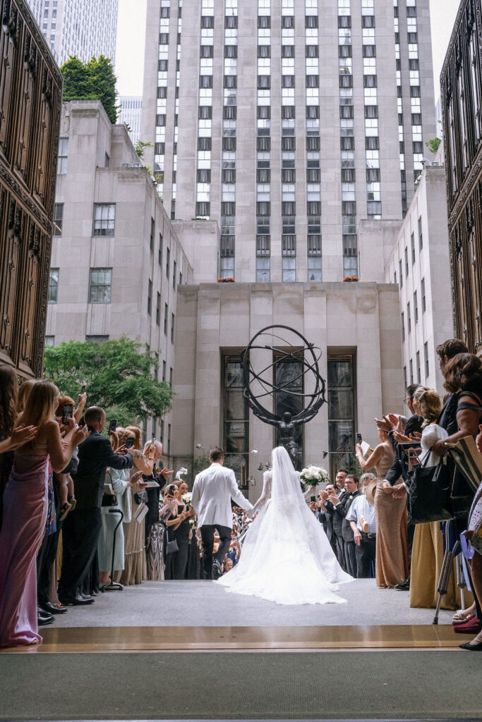 The bride and groom exit the cathedral hand-in-hand, greeted by cheering guests with the Atlas statue and NYC skyline in the background.