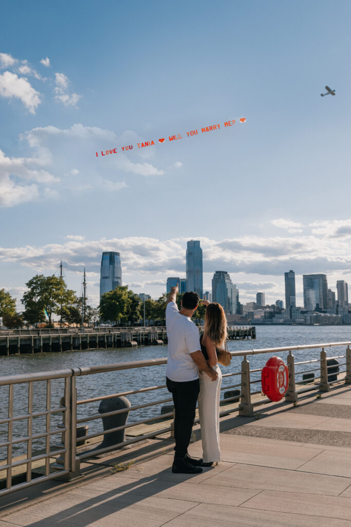 Bride and groom embracing under a clear blue sky with a private plane proposal banner flying overhead in NYC.