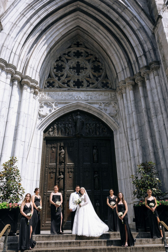 The bride and groom pose with their bridesmaids in elegant black dresses on the grand steps of Saint Patricks Cathedral NYC.