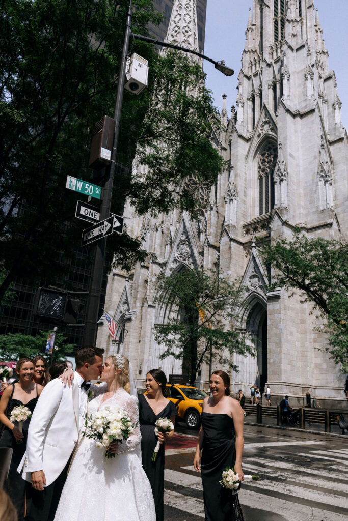 The bride and groom share a kiss outside the NYC cathedral, surrounded by their bridal party in black dresses and the city’s iconic yellow taxi in the background.