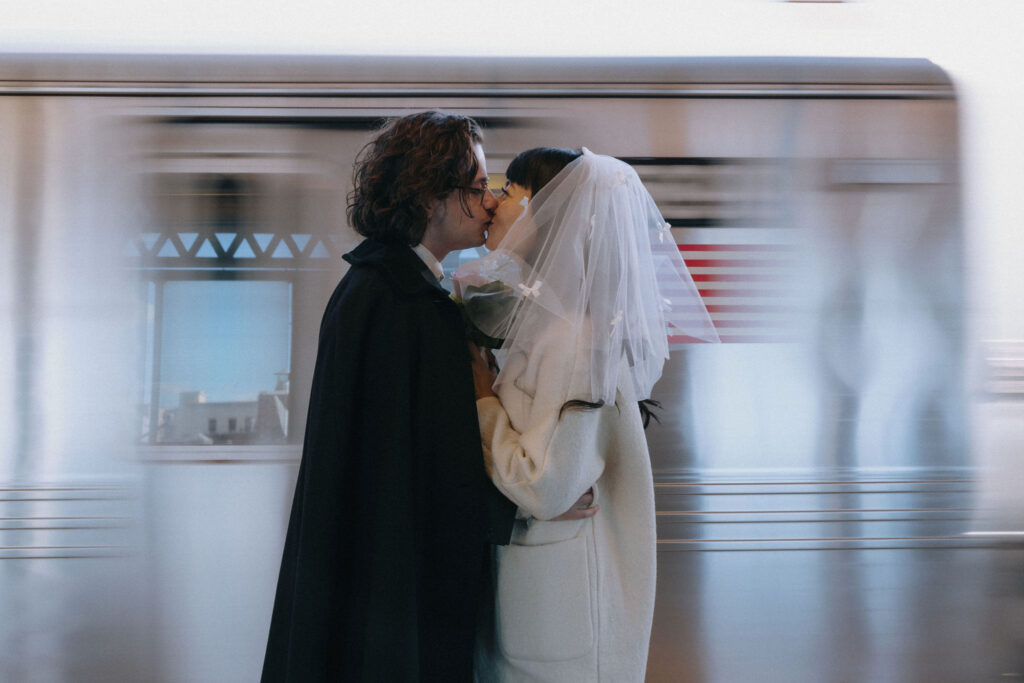 A bride and groom share a romantic kiss on a subway platform in New York City. The bride wears a white coat and veil adorned with delicate bows, while the groom dons a black cape. A train speeds by in the background, capturing the motion and energy of the city.