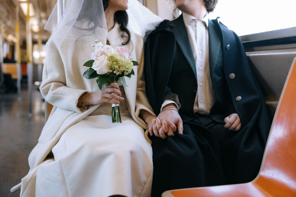 Close-up of a newlywed couple seated on a subway train, holding hands. The bride holds a bouquet of pale pink roses, white hydrangeas, and greenery, while wearing a white coat and veil. The groom is dressed in a suit with a dark overcoat, their connection emphasized through a tender gesture