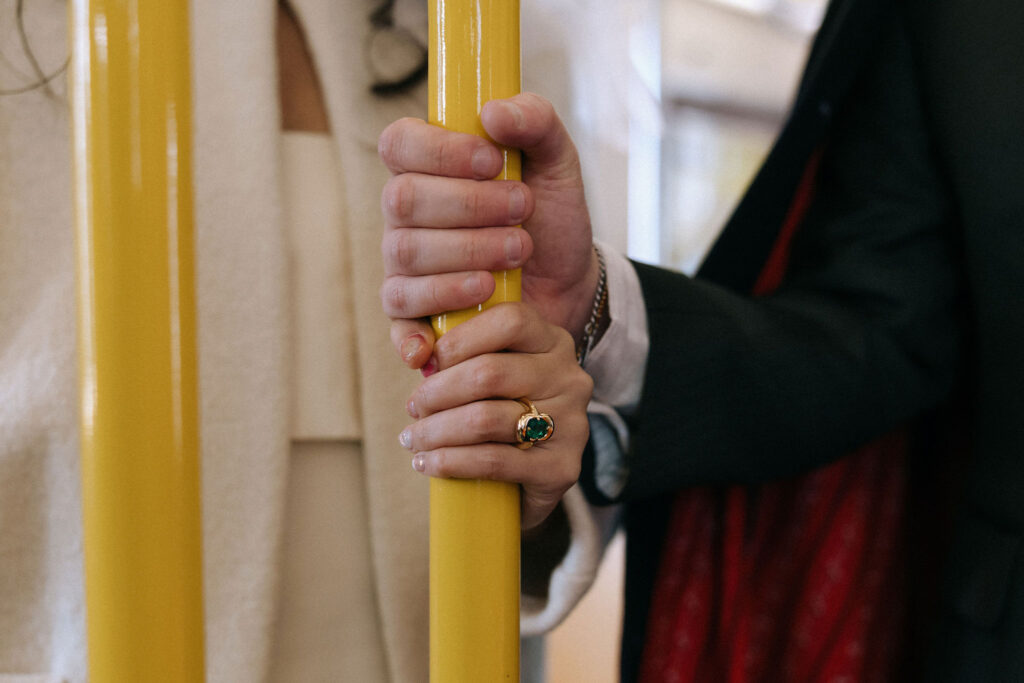 Close-up of a newlywed couple’s hands clasped together on a yellow subway pole. The bride wears an emerald ring, and the groom’s hand gently overlaps hers, symbolizing unity and support