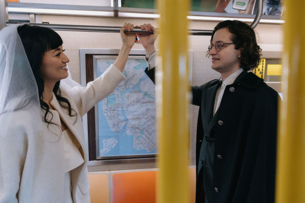 Newlyweds share a playful moment on a New York City subway, holding onto the same bar while gazing at each other. The bride beams with joy, her veil cascading over her shoulder, as the groom smiles softly, framed by the subway map in the background