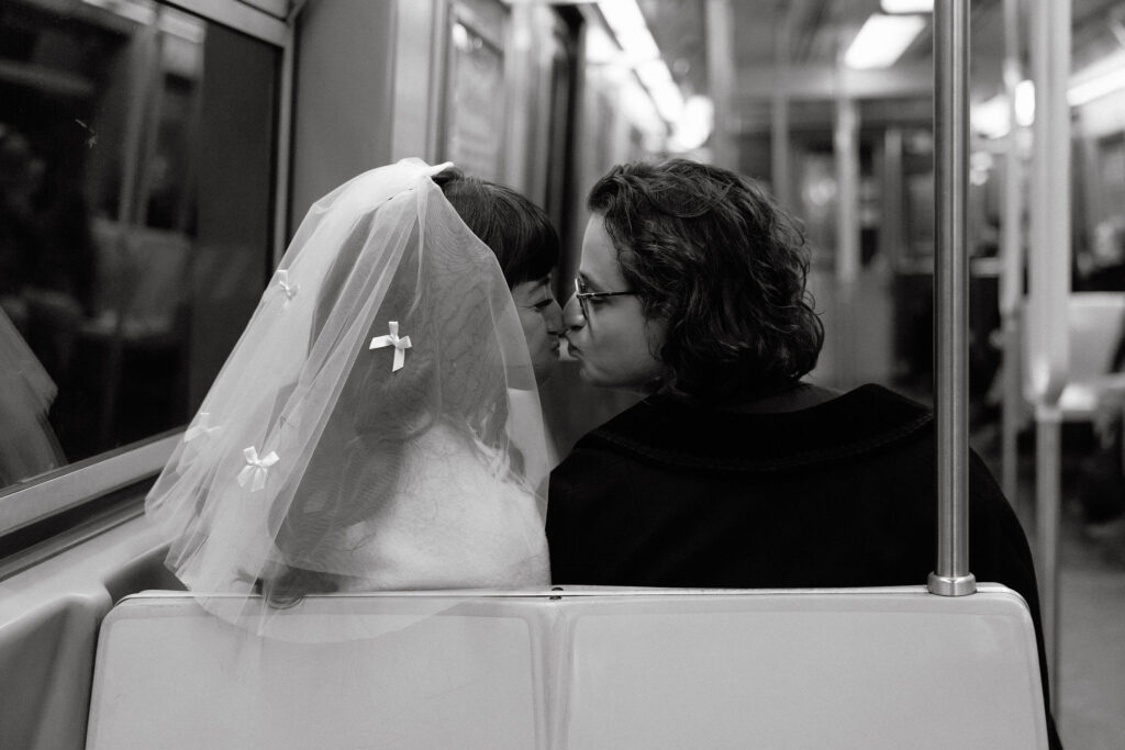 Black-and-white photo of a newlywed couple sharing a tender kiss on a New York City subway seat. The bride’s delicate veil, adorned with small bows, cascades softly, while the groom leans in, creating a moment of intimacy amidst the bustling urban setting