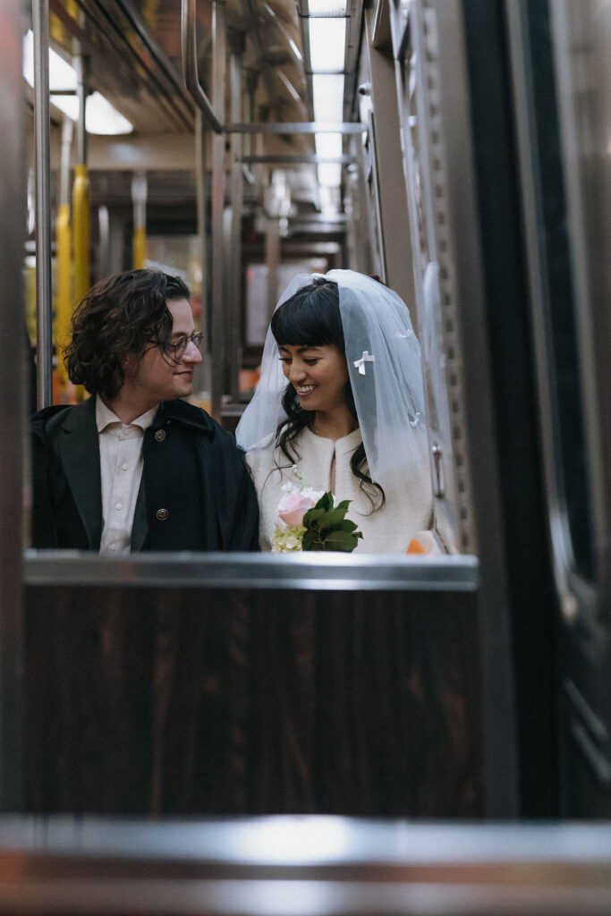 A candid moment of a newlywed couple sitting together on an NYC subway. The bride, wearing a veil adorned with small bows, smiles brightly while holding a delicate bouquet of pink roses and greenery. The groom gazes at her with a loving expression, dressed in a suit and coat, capturing a romantic yet urban vibe