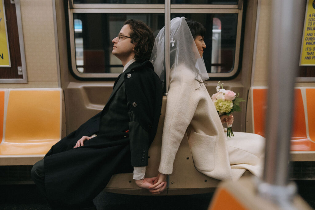 A candid photo of a newlywed couple on an NYC subway train. The groom leans back against the bride, both sitting on adjacent seats with their backs touching, holding hands, and smiling softly. The bride holds a bouquet of pink and white flowers, her veil adding a whimsical touch against the subway's bright orange and yellow seats.