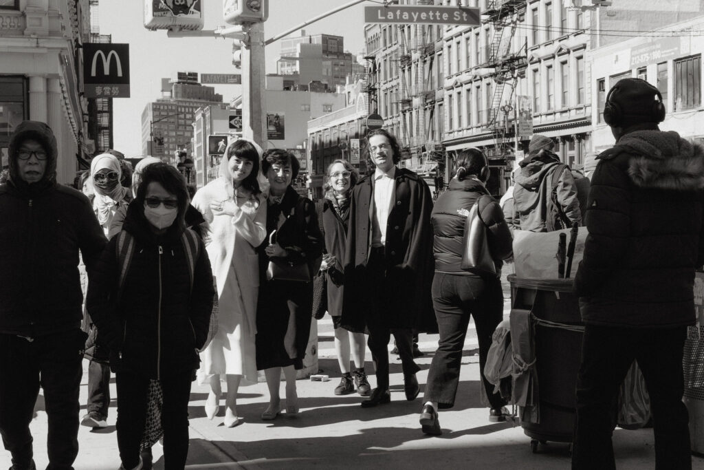 A black-and-white candid photo of a wedding group walking on a bustling city street near Lafayette Street. The bride is dressed in a white coat, smiling warmly, while the groom wears a dark coat. Family and friends accompany them, blending into the vibrant city crowd