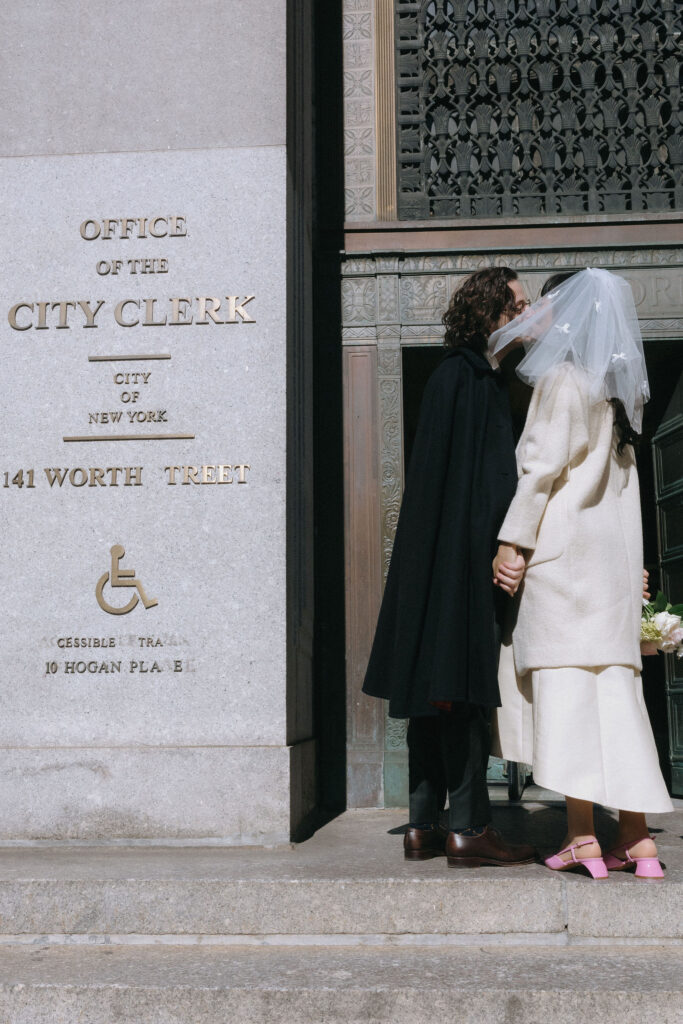 A newlywed couple shares a kiss outside the New York City Clerk's Office at 141 Worth Street. The bride wears a white coat with a veil adorned with delicate bows and pink shoes, while the groom is dressed in a dark suit with a cape. They hold hands in front of the building’s ornate details and signage.