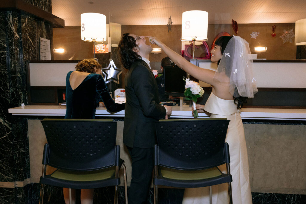 A playful moment at a city hall marriage bureau as the bride, holding a bouquet, feeds a piece of food to the groom while he leans back laughing. In the background, a woman in a velvet dress signs papers at the counter under warm lighting, with decorations adding charm to the scene.