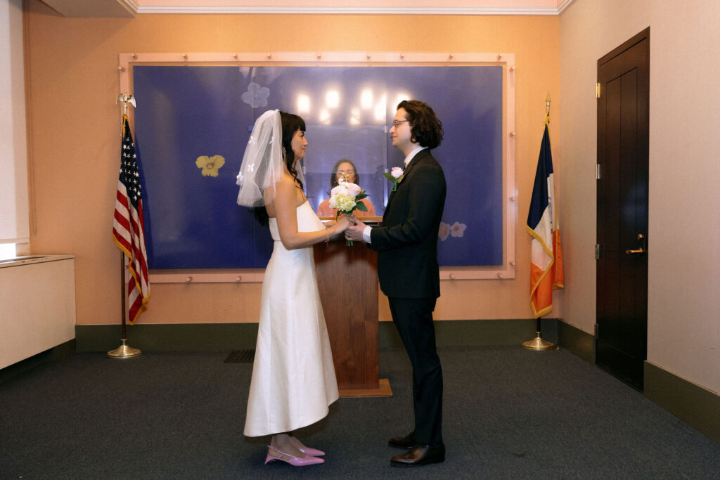 A bride and groom stand face to face, holding hands, during their wedding ceremony at a city hall. The bride wears a simple white dress and veil, while the groom is in a classic suit. Behind them, the officiant stands at a wooden podium, with an American flag and a New York City flag framing the room.