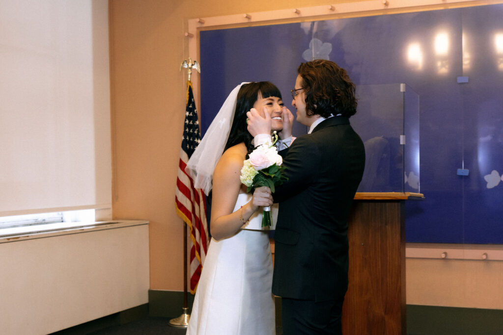 A groom tenderly cups the bride's face as she smiles brightly during their wedding ceremony. She holds a bouquet of pink and white flowers, standing near an American flag and a wooden podium