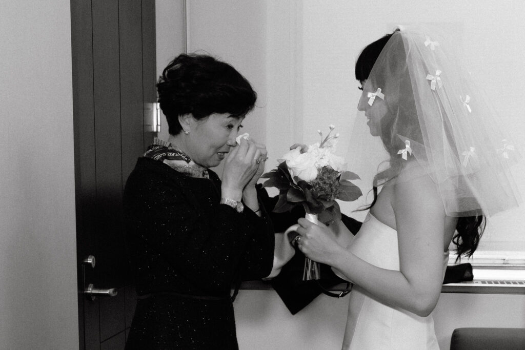 A heartfelt moment between a bride and her mother as tears of joy are shared. The bride, holding her bouquet, wears a veil adorned with delicate bows, while her mother wipes her eyes in an emotional exchange