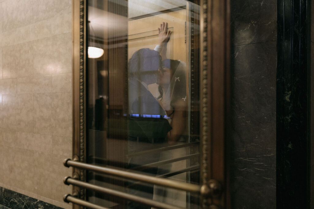 Romantic reflection of a couple kissing through a vintage glass elevator door. The warm tones of the wooden frame and marble walls add a timeless elegance to the intimate moment.