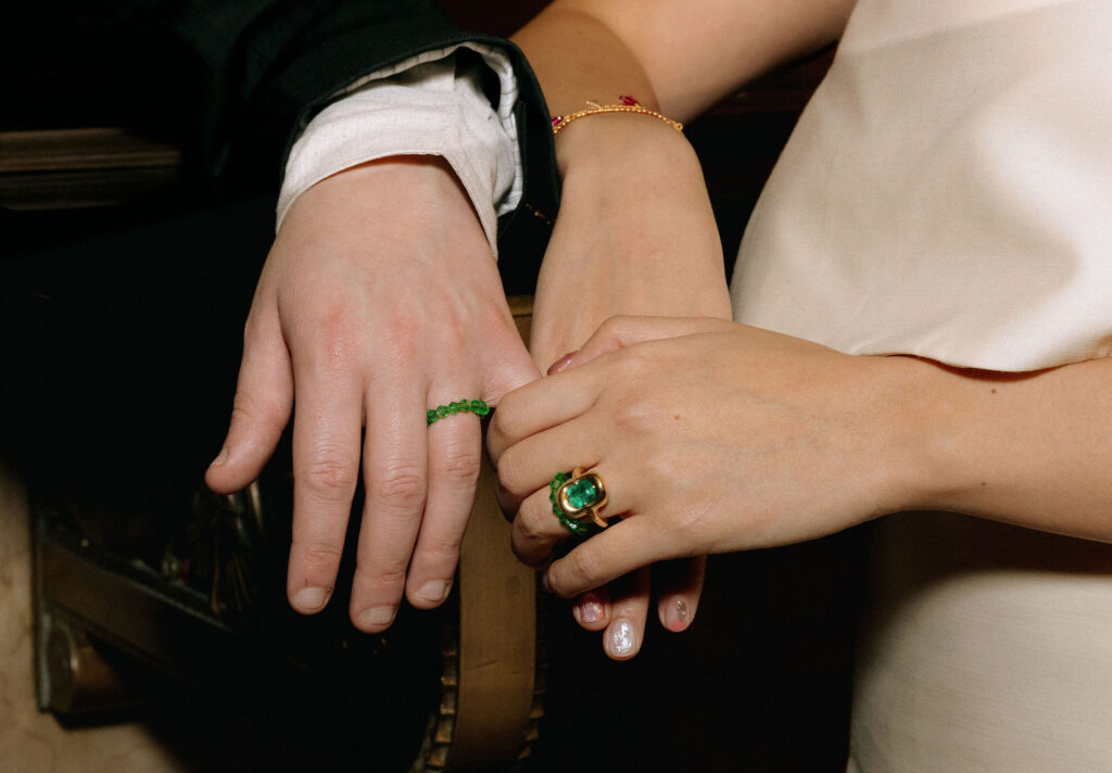 Close-up of a couple’s hands adorned with green rings. The bride’s hand features an elegant green gemstone ring with gold detailing, while the groom wears a simple green beaded ring. The bride also wears a delicate gold bracelet with red accents