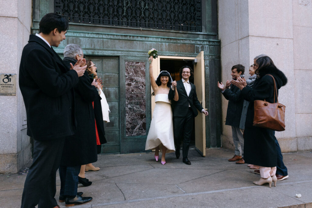 The newlywed couple jubilantly exits the building, hand in hand, with the bride raising her bouquet in celebration. Friends and family stand on either side, clapping and cheering in joy