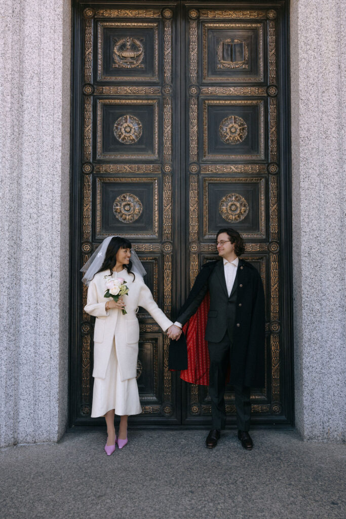 A wedding couple stands hand-in-hand in front of an ornate, intricately designed bronze door framed by stone pillars. The bride wears a cream outfit with a veil, holding a pastel bouquet, while the groom wears a suit and cape lined with red fabric, glancing lovingly at each other