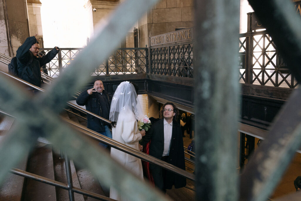 A candid moment of a bride and groom walking up subway stairs in NYC, captured through a metal railing. The groom gazes up at the bride, while onlookers in the background cheer and pose enthusiastically. The subway entrance sign adds a quintessential NYC charm to the scene