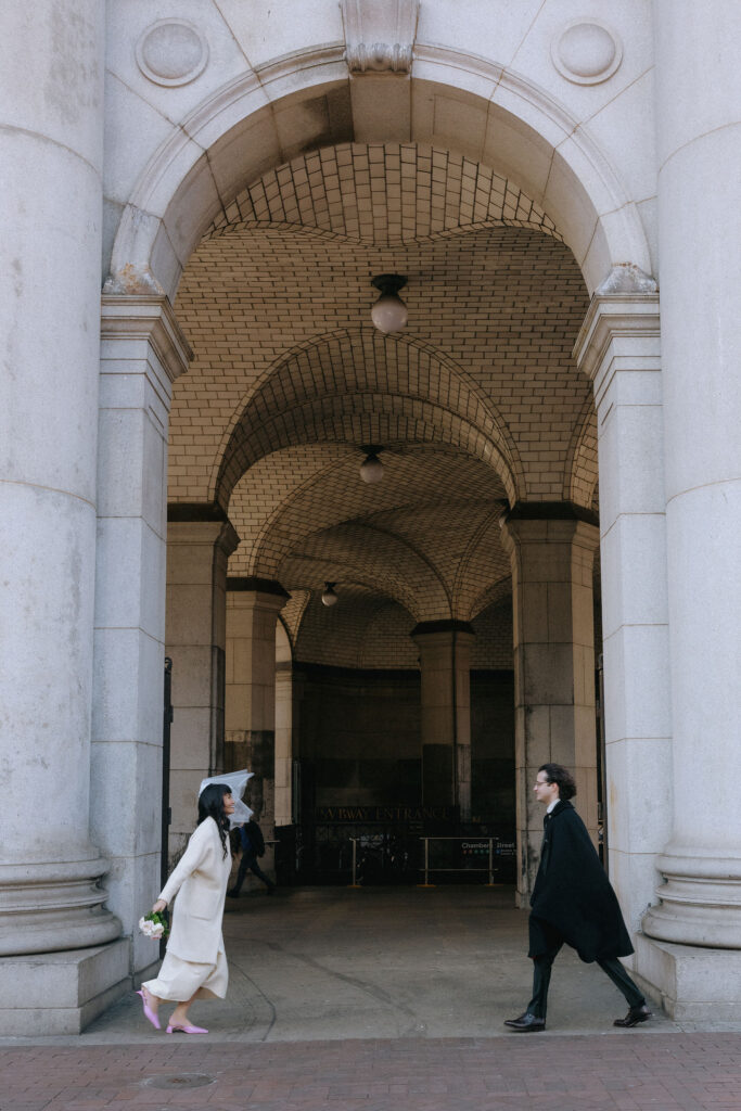 The bride and groom walk towards each other under the elegant tiled arches of a historic NYC building. The bride holds a bouquet and wears a flowing white outfit paired with pink heels, while the groom dons a black cape, creating a cinematic and timeless moment