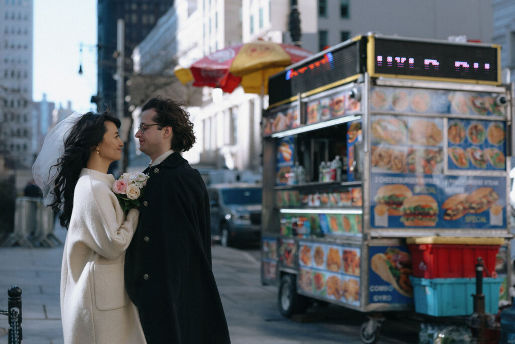 A bride and groom share a loving gaze in front of a vibrant NYC food cart. The bride holds a bouquet of pastel roses, and the groom wears a black cape. The backdrop features the energy of the city, with bright food cart signage and urban architecture.