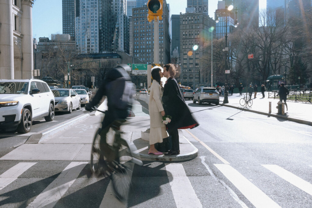 Newlyweds share a kiss at a busy NYC crosswalk, surrounded by urban life and motion, with a cyclist passing in the foreground