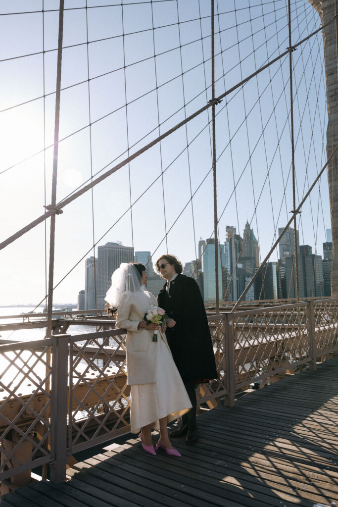 Newlywed couple sharing an intimate moment on the Brooklyn Bridge, with the Manhattan skyline and morning sun creating a romantic atmosphere.