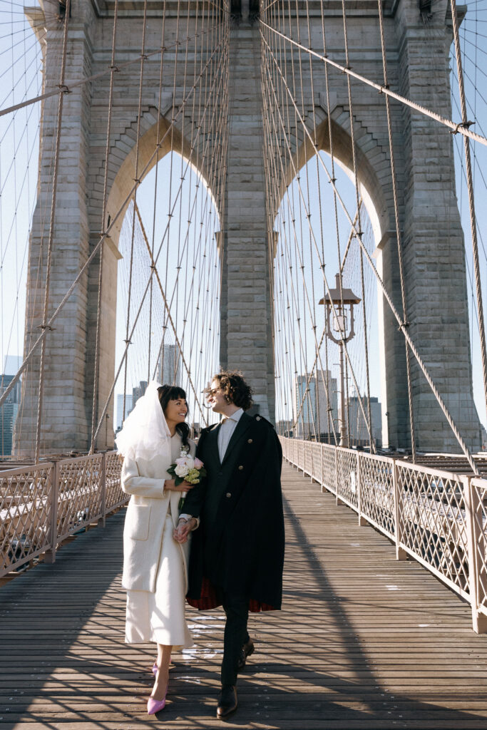 Smiling newlywed couple walking hand-in-hand on Brooklyn Bridge, framed by its iconic stone arches and suspension cables, with the city skyline in the background.