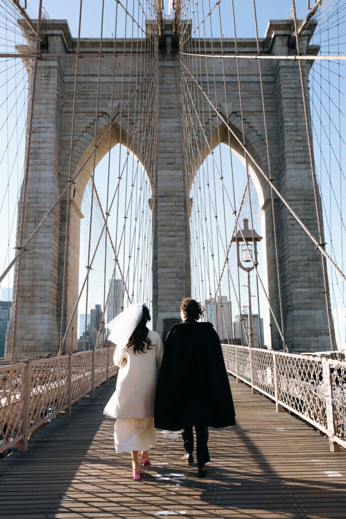 Newlywed couple walking hand-in-hand on the iconic Brooklyn Bridge, framed by its soaring arches and intricate cables under a bright blue sky.