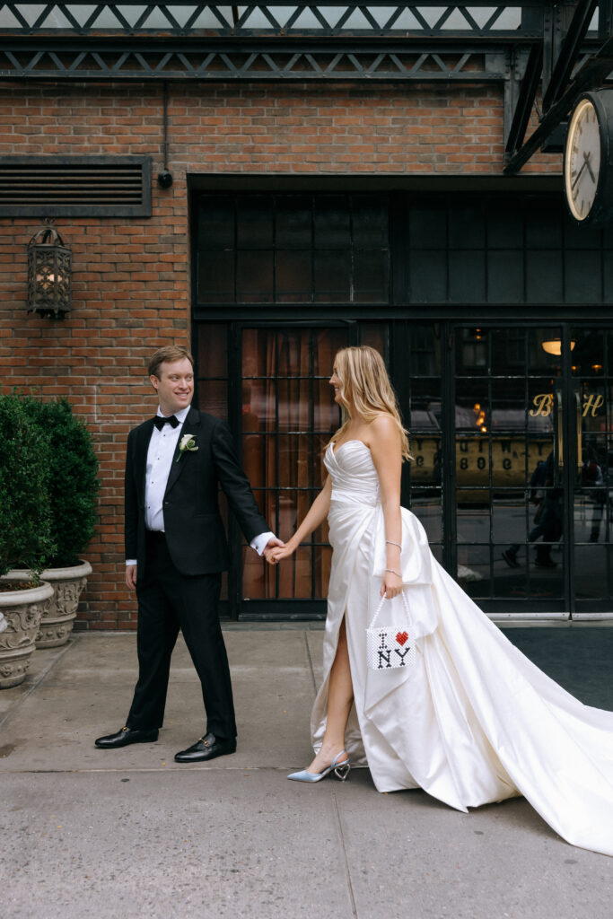 Stylish bride and groom walking hand in hand outside the Bowery Hotel, with the bride showcasing a custom 'I ❤️ NY' bag and elegant satin gown.