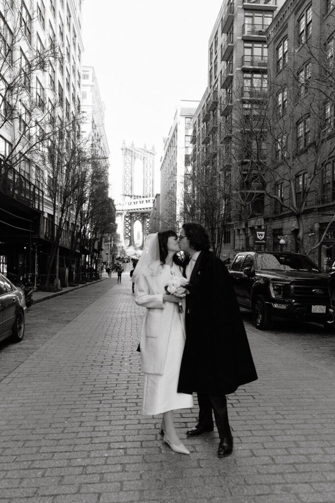 Newlywed couple sharing a kiss on a cobblestone street in DUMBO, Brooklyn, with the iconic Manhattan Bridge in the background