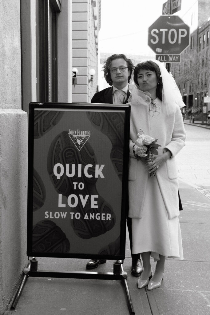 A bride and groom standing behind a sign reading 'Quick to Love, Slow to Anger,' on a city street with a stop sign in the background