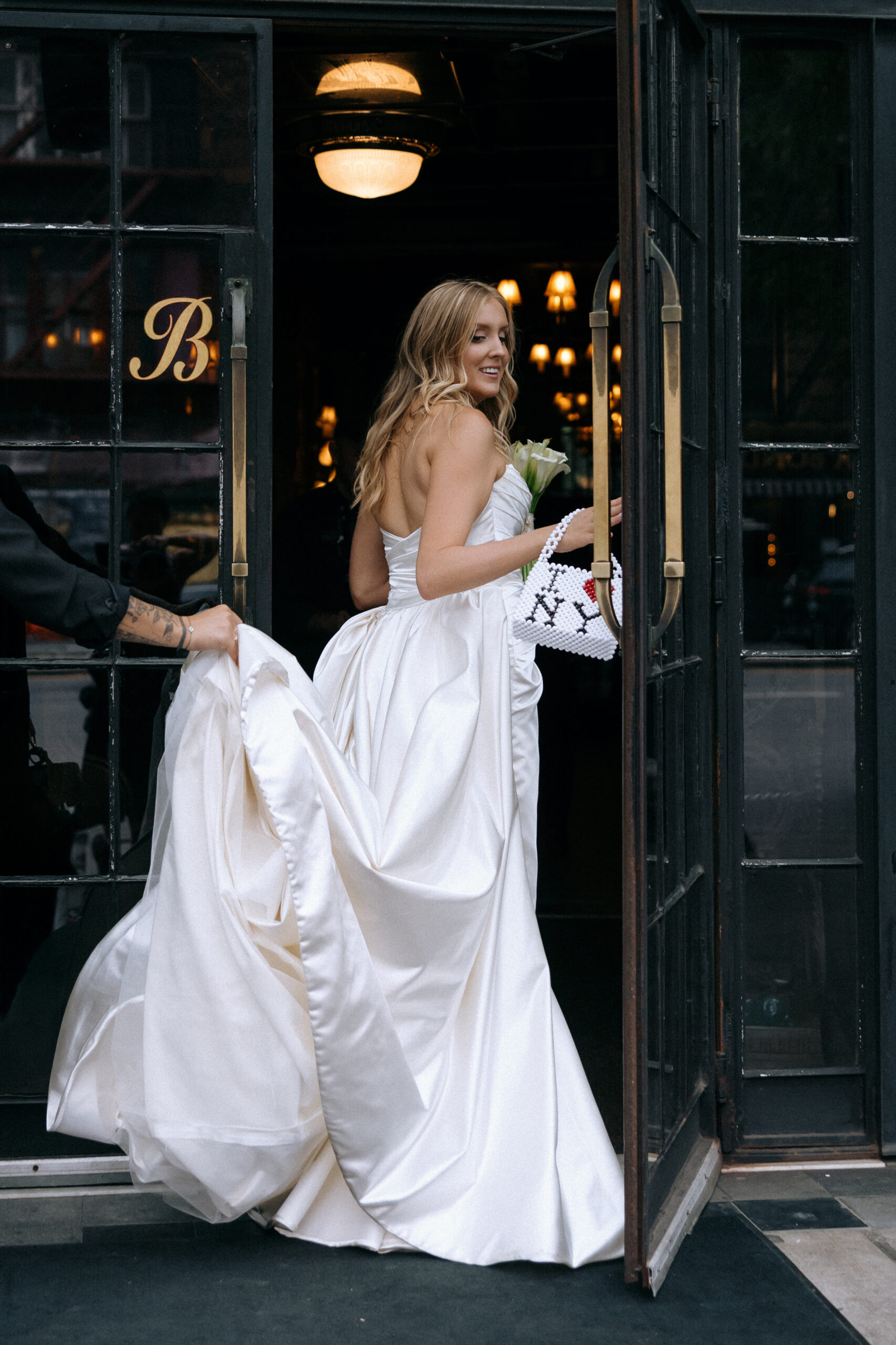 Smiling bride in a strapless satin gown holding an 'I ❤️ NY' beaded bag and her train as she steps into the Bowery Hotel.