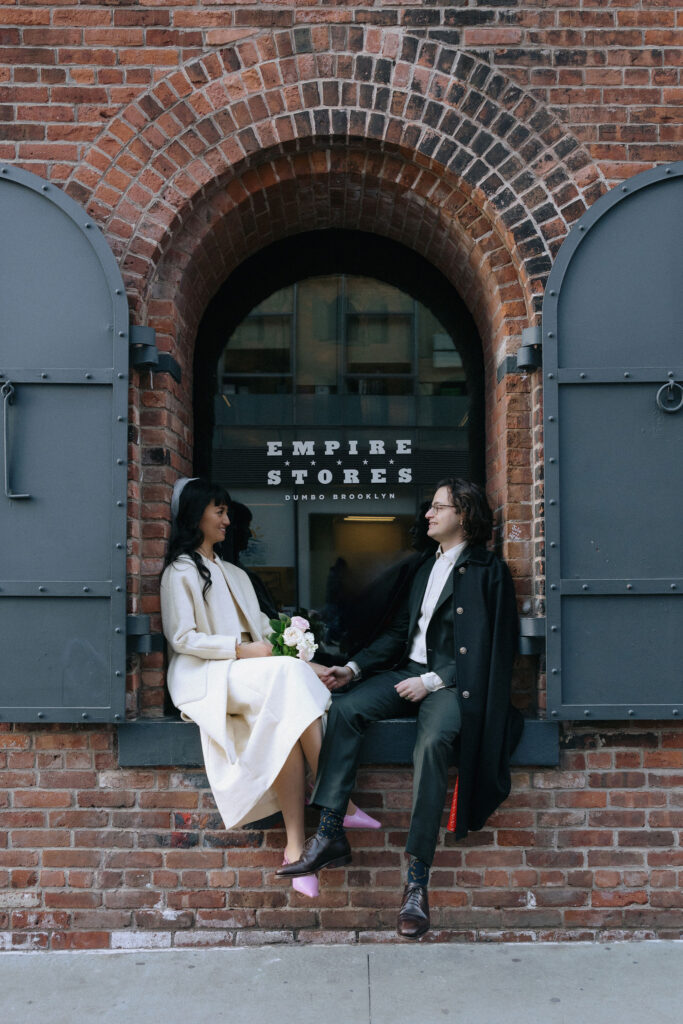 A bride and groom seated on a windowsill at Empire Stores in DUMBO, Brooklyn, holding hands and smiling at each other against a rustic brick backdrop