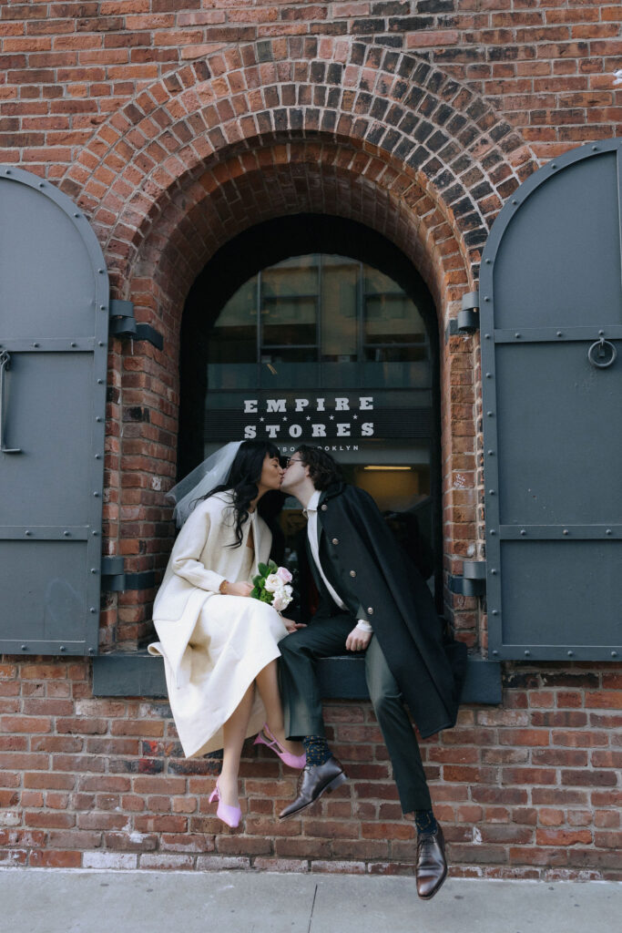 A couple shares a kiss while sitting on a windowsill at the Empire Stores in Brooklyn, with a romantic backdrop of rustic red bricks and industrial shutters.