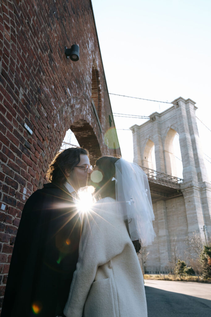 A romantic kiss captured between a bride and groom with the Brooklyn Bridge and sunlight shining through their embrace, framed by a red brick archway.