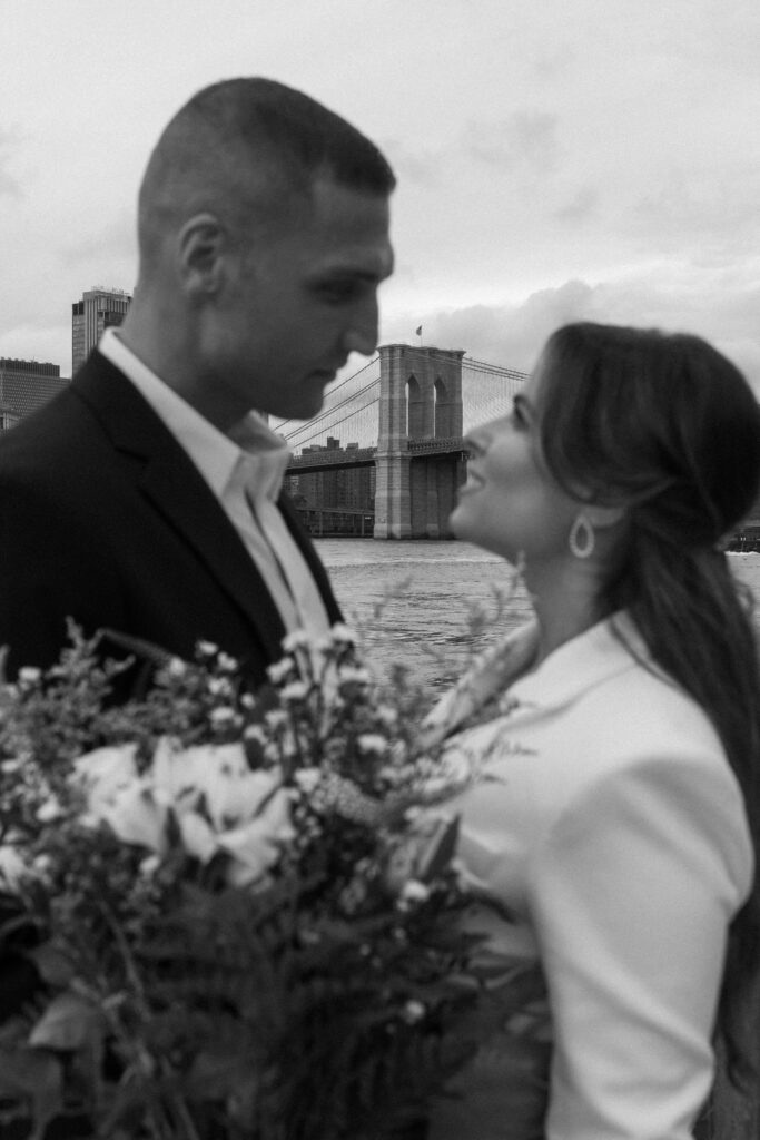 Black and white photo of a couple embracing near the Brooklyn Bridge during their NYC wedding.