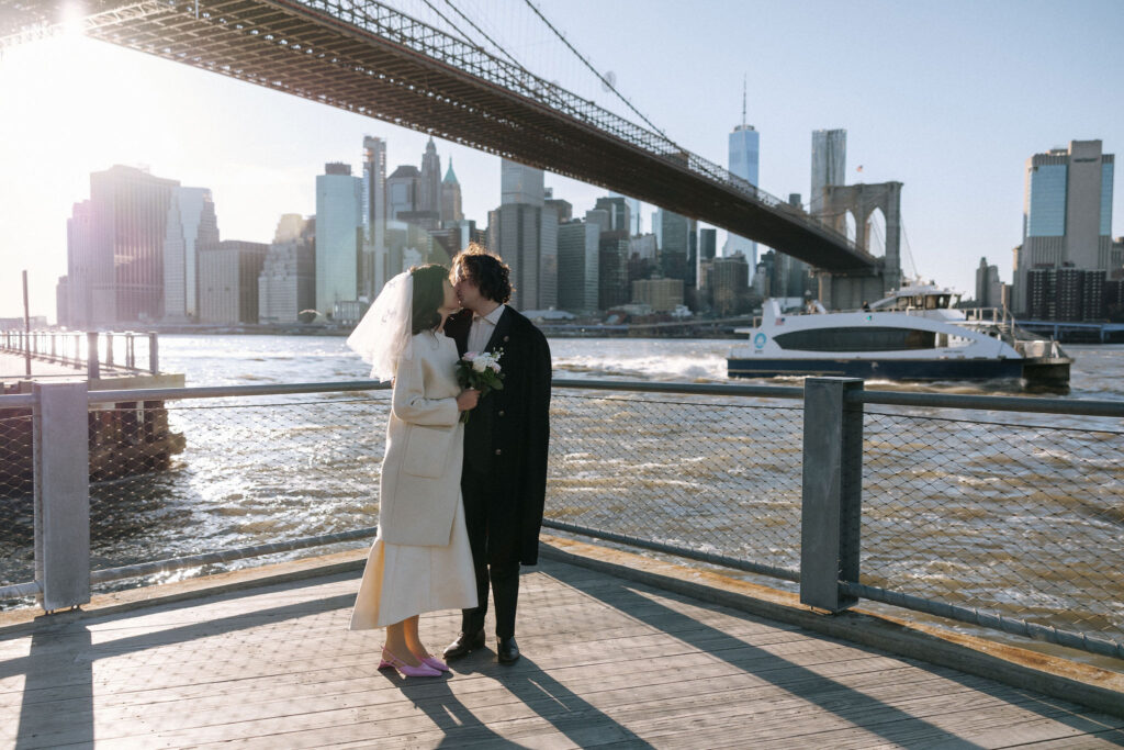 A bride and groom share a romantic kiss on the waterfront with the Brooklyn Bridge and NYC skyline glowing in the sunlight.