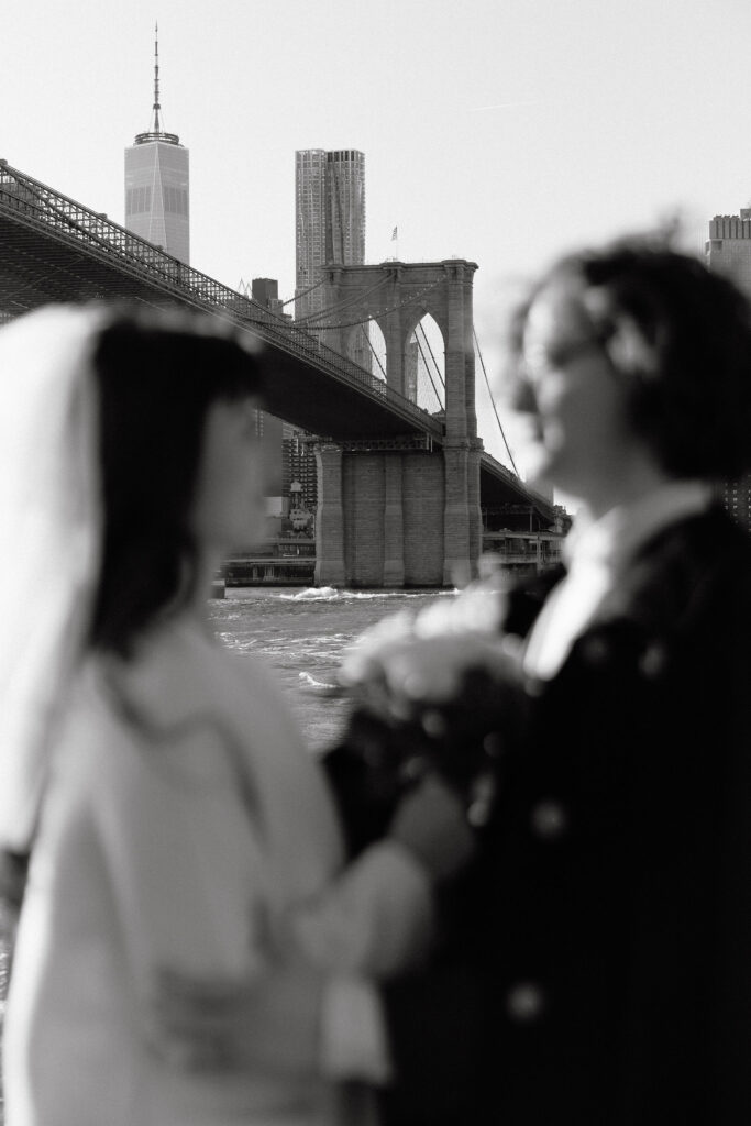 Black and white image of the Brooklyn Bridge with a blurred silhouette of a bride and groom in the foreground, holding hands