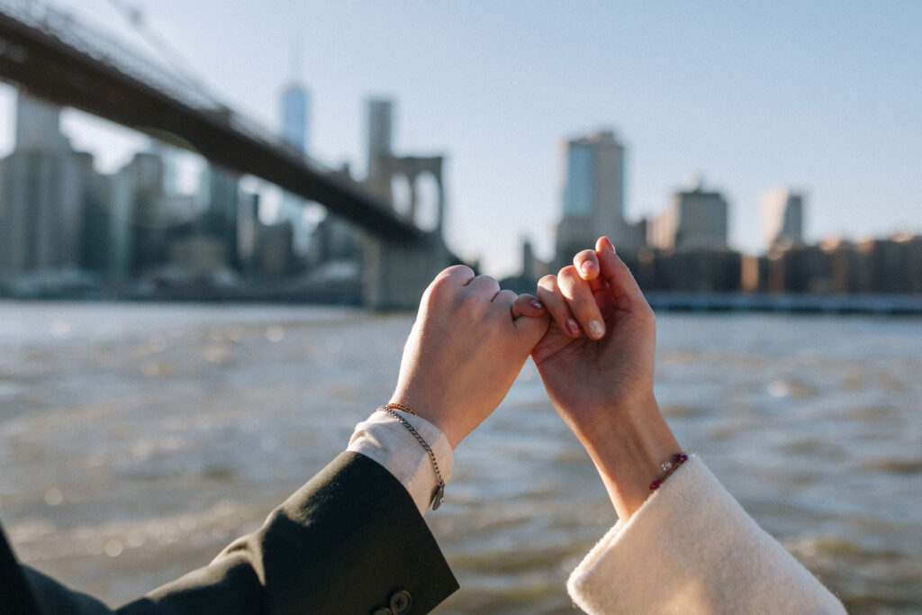 Close-up of a couple's hands linking pinky fingers in a promise gesture, with the Brooklyn Bridge and the Manhattan skyline blurred in the background. Sunlight sparkles on the water.