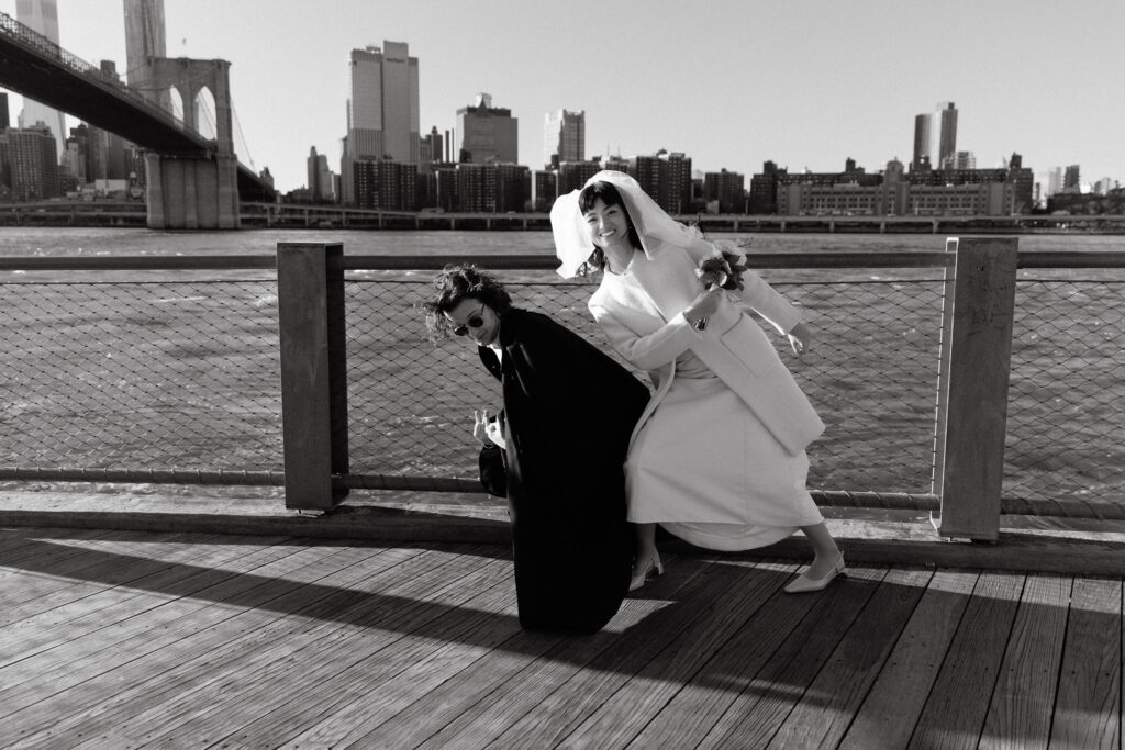 Black and white photo of a playful couple on a Brooklyn Bridge Park pier. The groom, in sunglasses and a dramatic black coat, strikes a dynamic pose, while the bride, holding a bouquet and wearing a veil, leans into the shot with a joyful smile. The Brooklyn Bridge and Manhattan skyline are in the background.