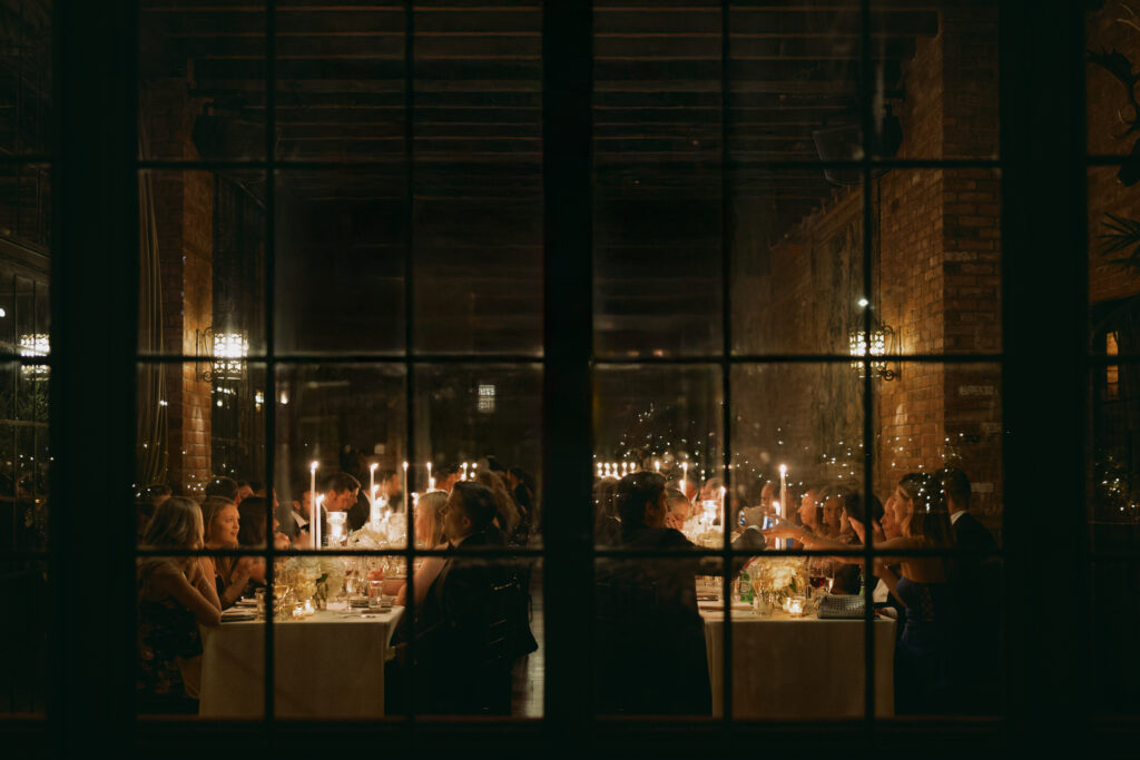 An intimate candlelit dinner reception at the Bowery Hotel, viewed through large paneled windows, capturing the warm glow and elegant atmosphere of the evening.